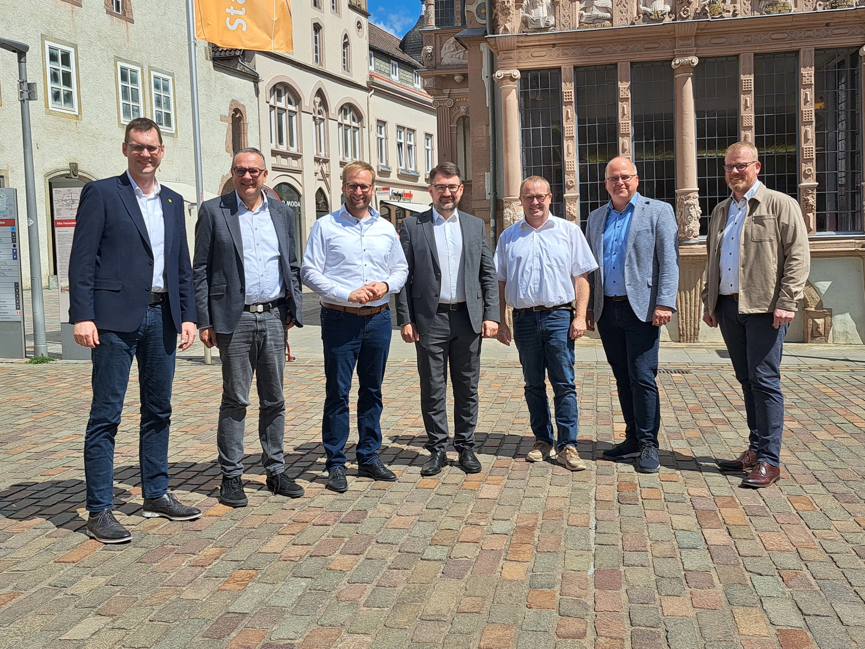 Sieben Männer stehen im Sonnenlicht vor dem Apothekenerker.