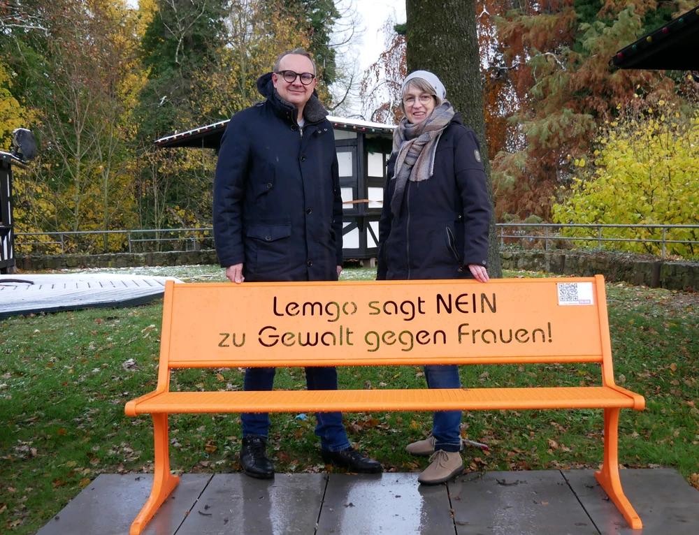 Im Vordergrund eine orange Bank. Dahinter zwei Personen.