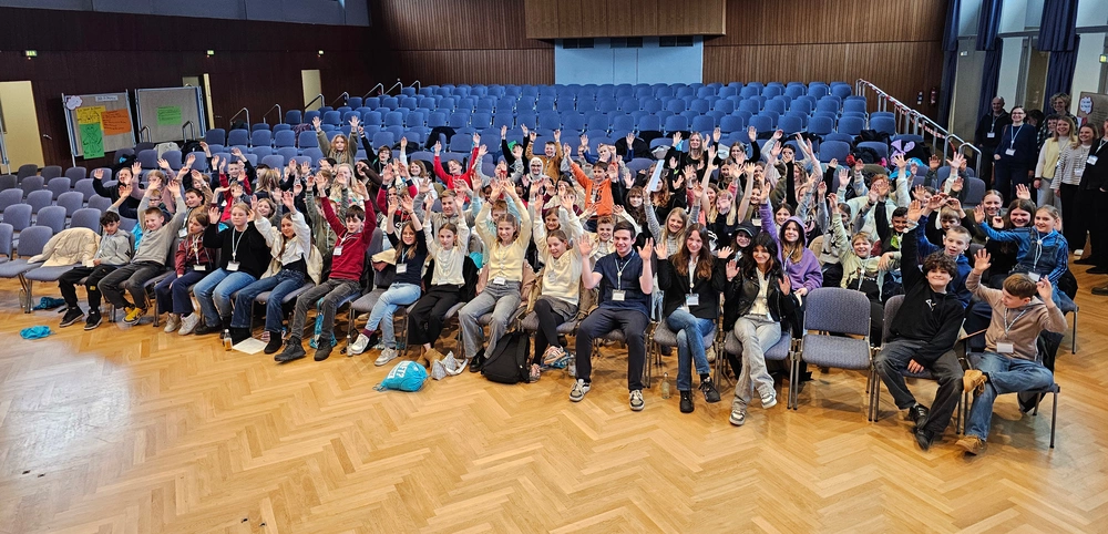 Eine große Gruppe von Kindern und Jugendlichen sitzt in einer Aula und hebt die Arme für das Foto.