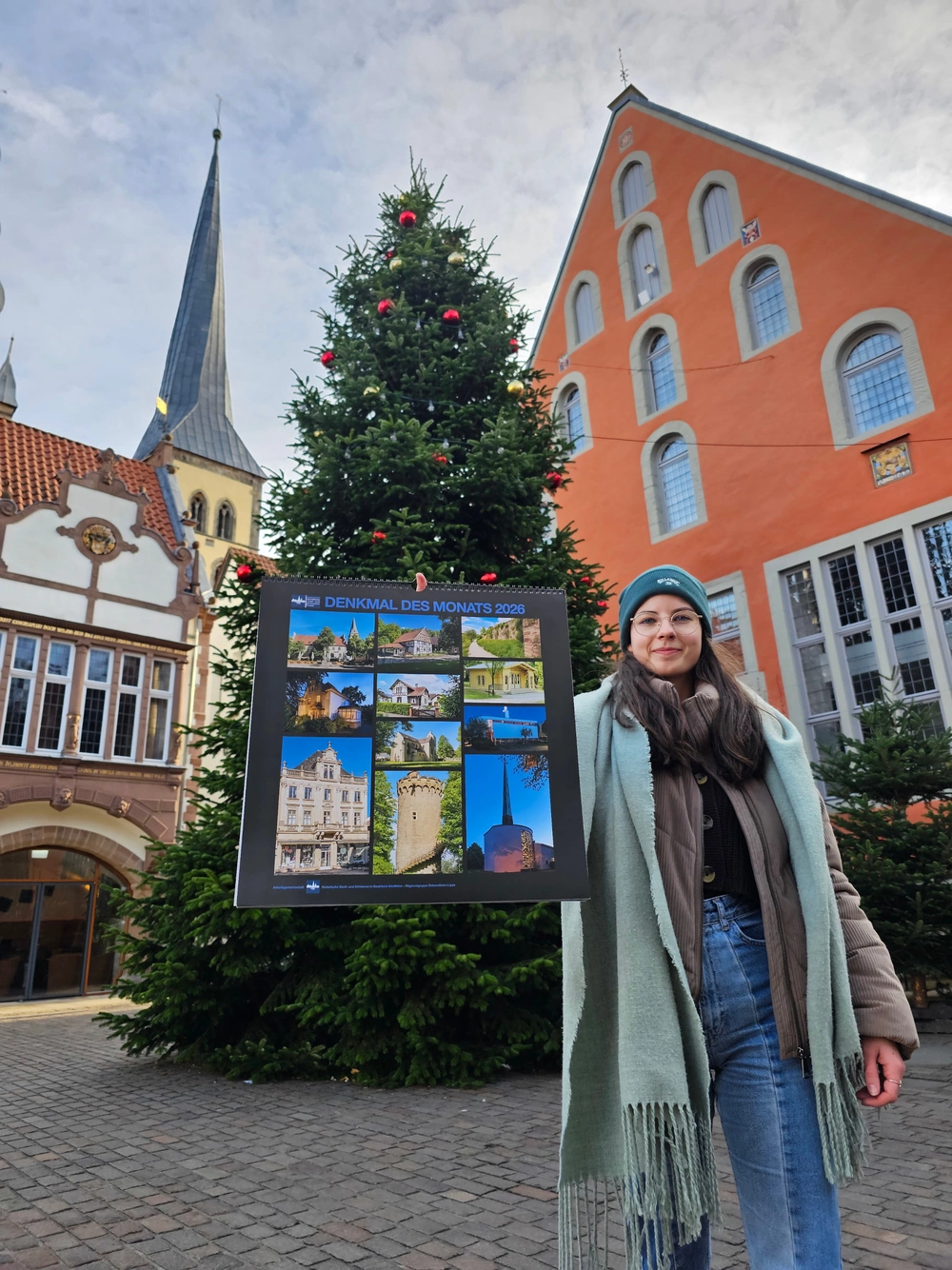 Eine junge Frau hält einen Jahreskalender in die Kamera. Im Hintergrund sind der Lemgoer Marktplatz, ein großer Tannenbaum, ein Teil des Rathauses, das Ballhaus und ein Turm der Kirche St. Nicolai zu sehen.