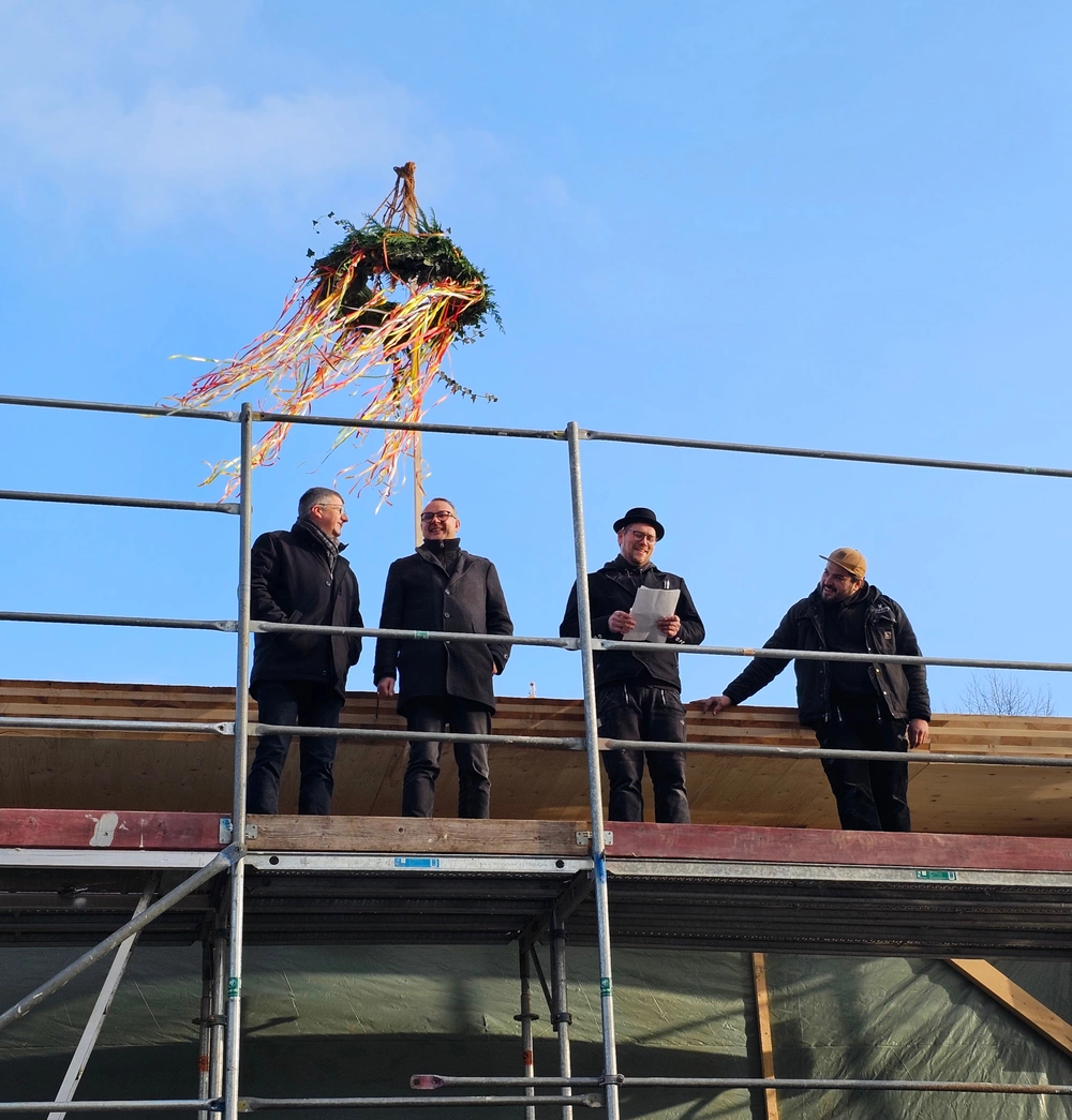 Vier Männer stehen auf einem Baugerüst um einen Rohbau. Hinter ihnen weht die Richtkrone im Wind.