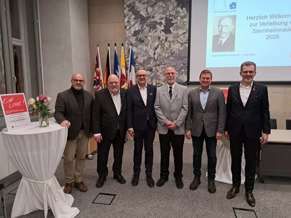 Sechs festlich gekleidete Männer stehen im Sitzungssaal des Rathauses nebeneinander.