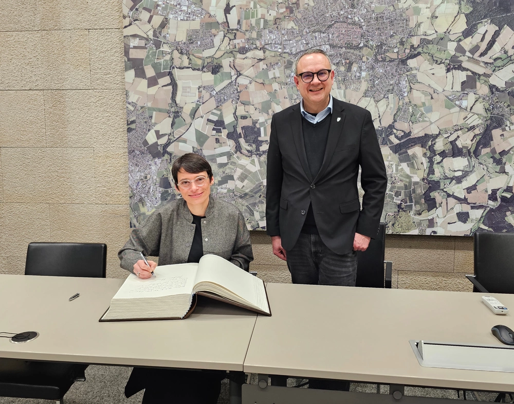 Eine Frau sitzt an einem Tisch und hat ein großes Buch vor sich liegen, ein Mann steht daneben.