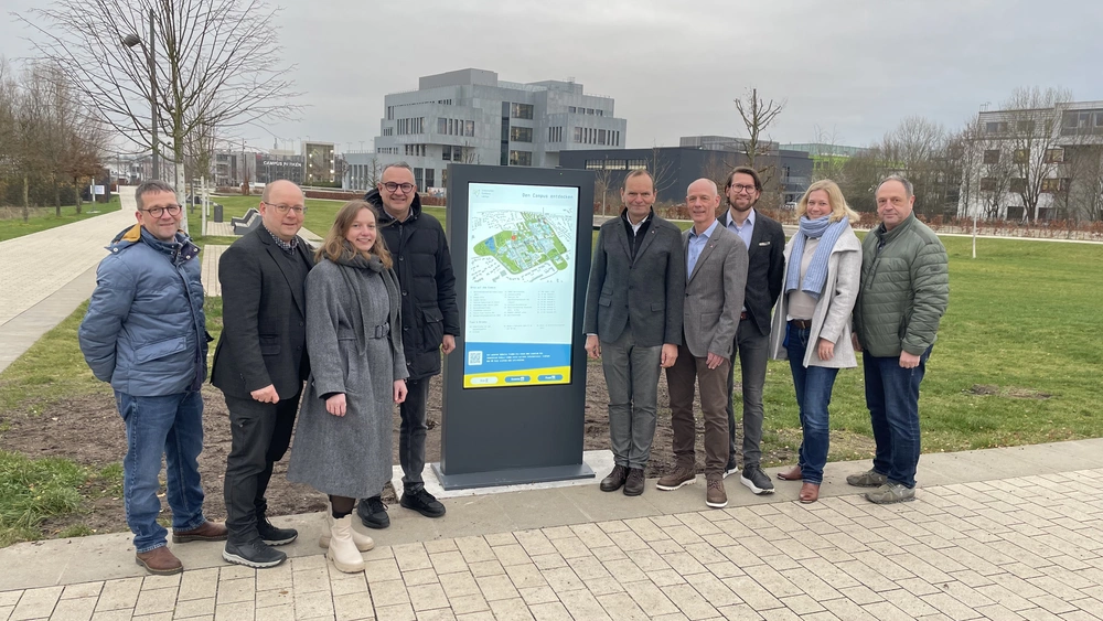 Neun Menschen stehen draußen neben einer Stele, die den Lageplan des Innovation Campus Lemgo zeigt. Im Hintergrund sieht man den Innovation Spin.