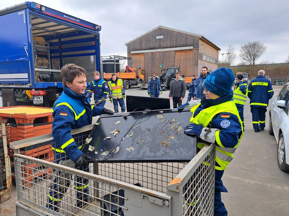 Zwei Jungen in THW-Uniform werfen einen dreckigen Fernseher in eine Gitterbox.