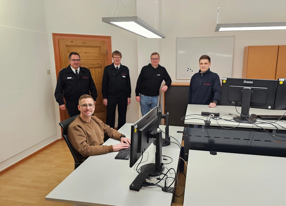 Fünf Männer sitzen und stehen um Schreibtische herum.