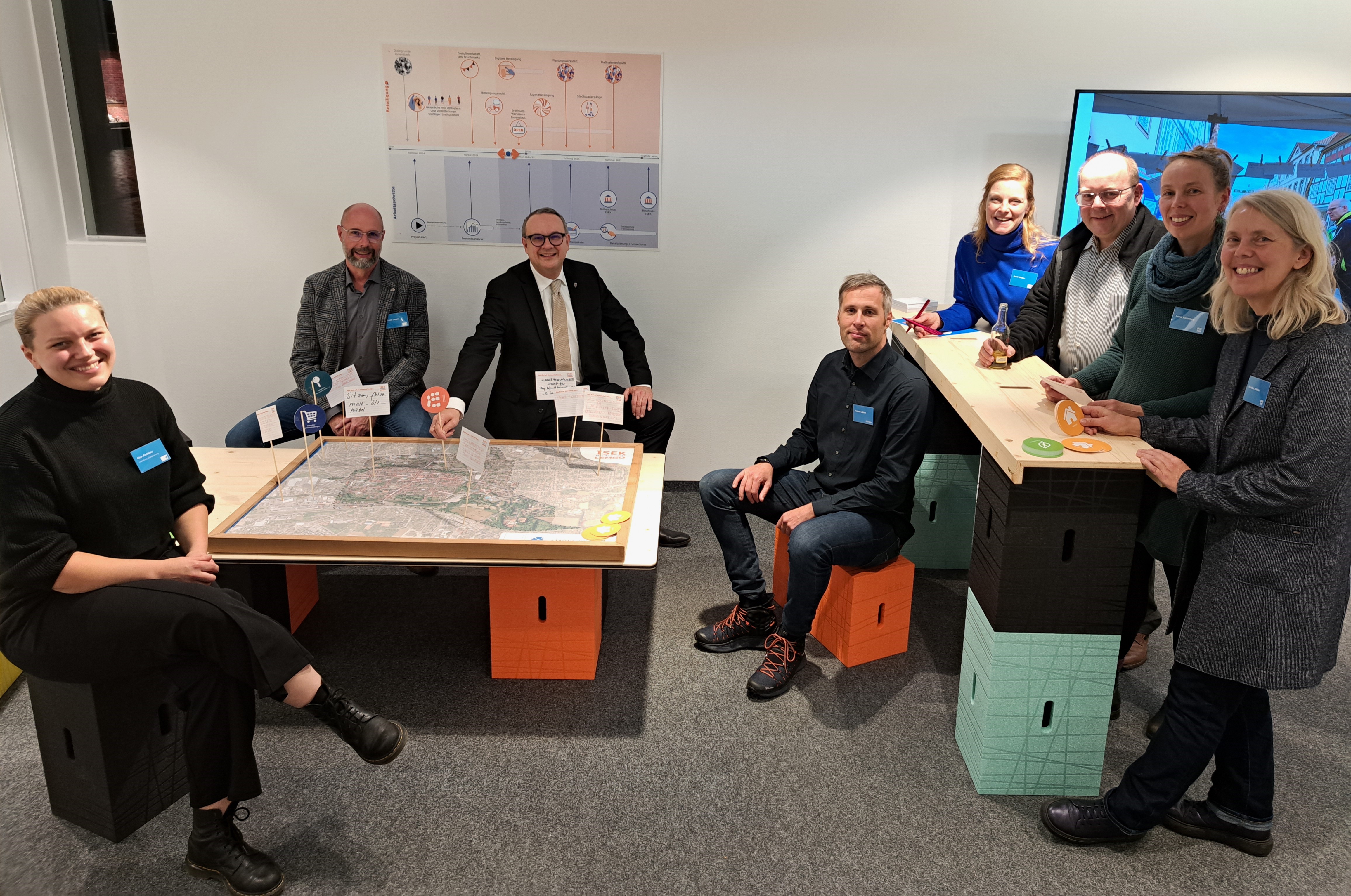 Acht Menschen sitzen und stehen im Gespräch beieinander. Auf einem niedrigen Tisch liegt ein Plan der Lemgoer Innenstadt, in en Holzstäbchen mit beschriebenen Zetteln gesteckt sind.