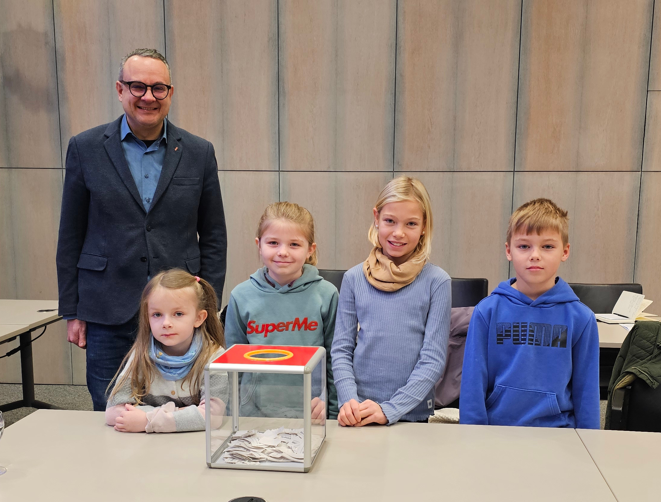 Ein Mann und vier Kinder stehen hinter einem Tisch, auf dem ein dursichtiger Würfel mit Papierschnipseln liegt.