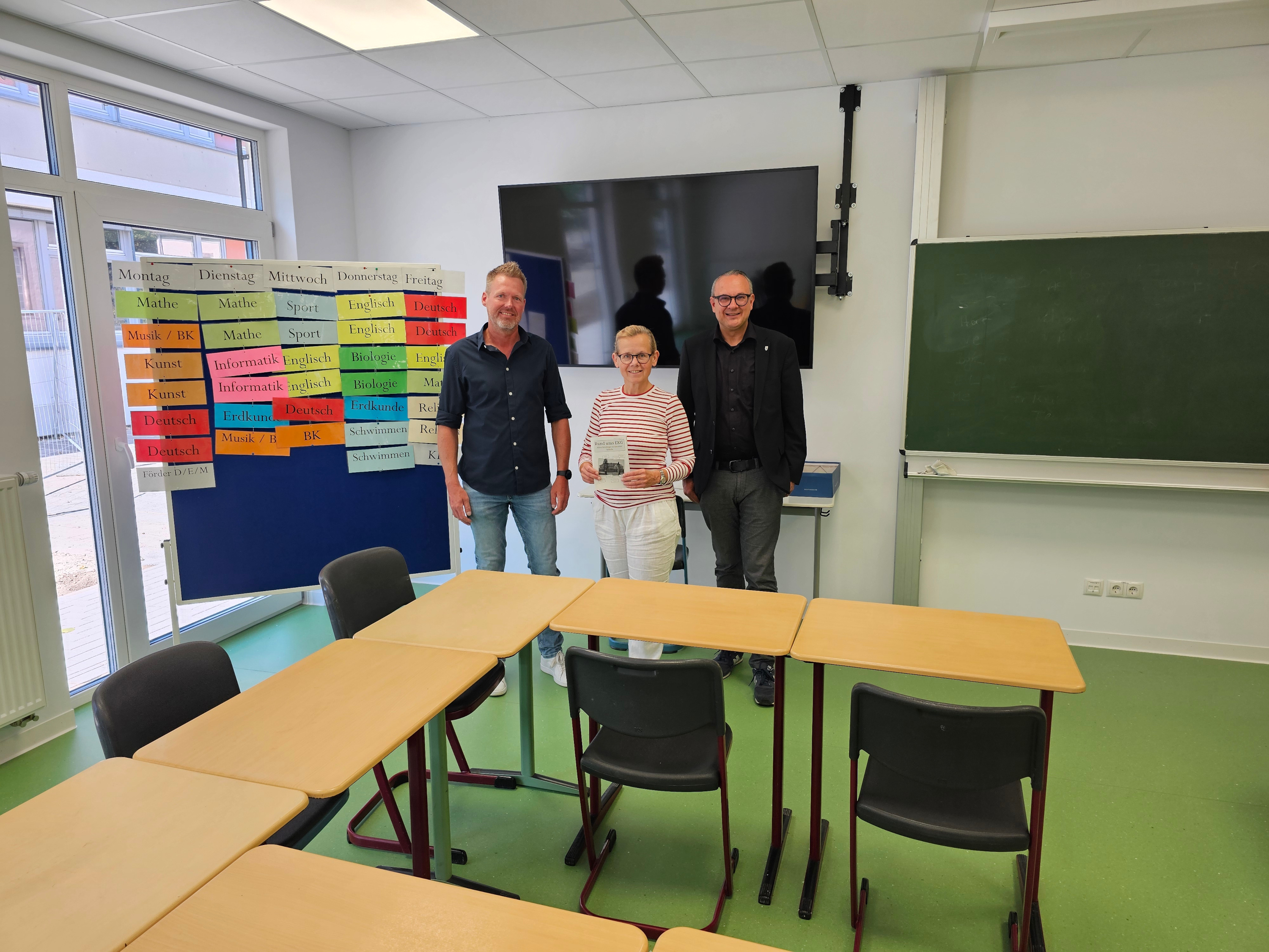 Drei Personen stehen in einem Klassenraum vor einem Stundenplan, einem Bildschirm und einer Tafel.