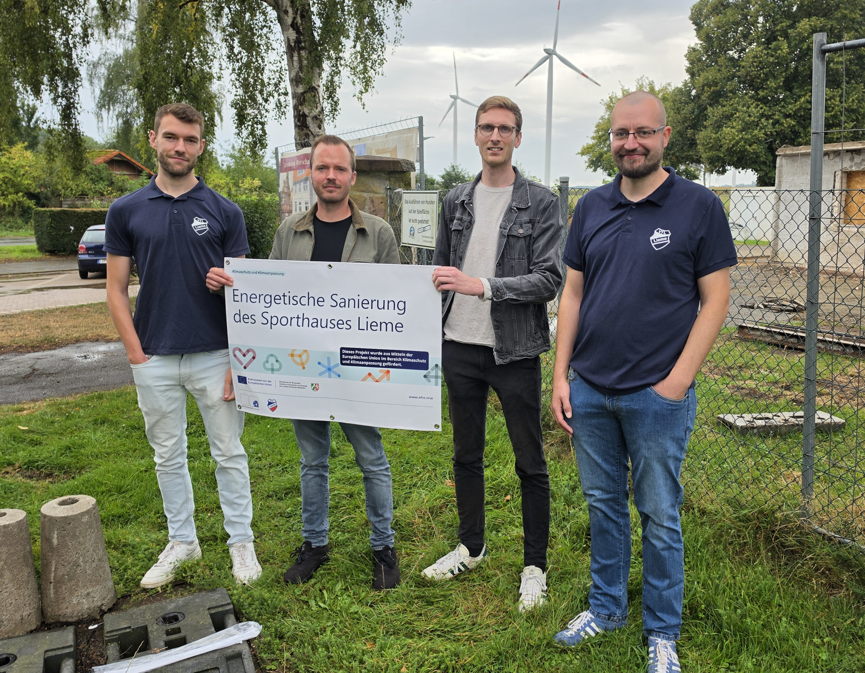 Vier Männer stehen vor einer Baustelle und halten ein Banner in der Hand.
