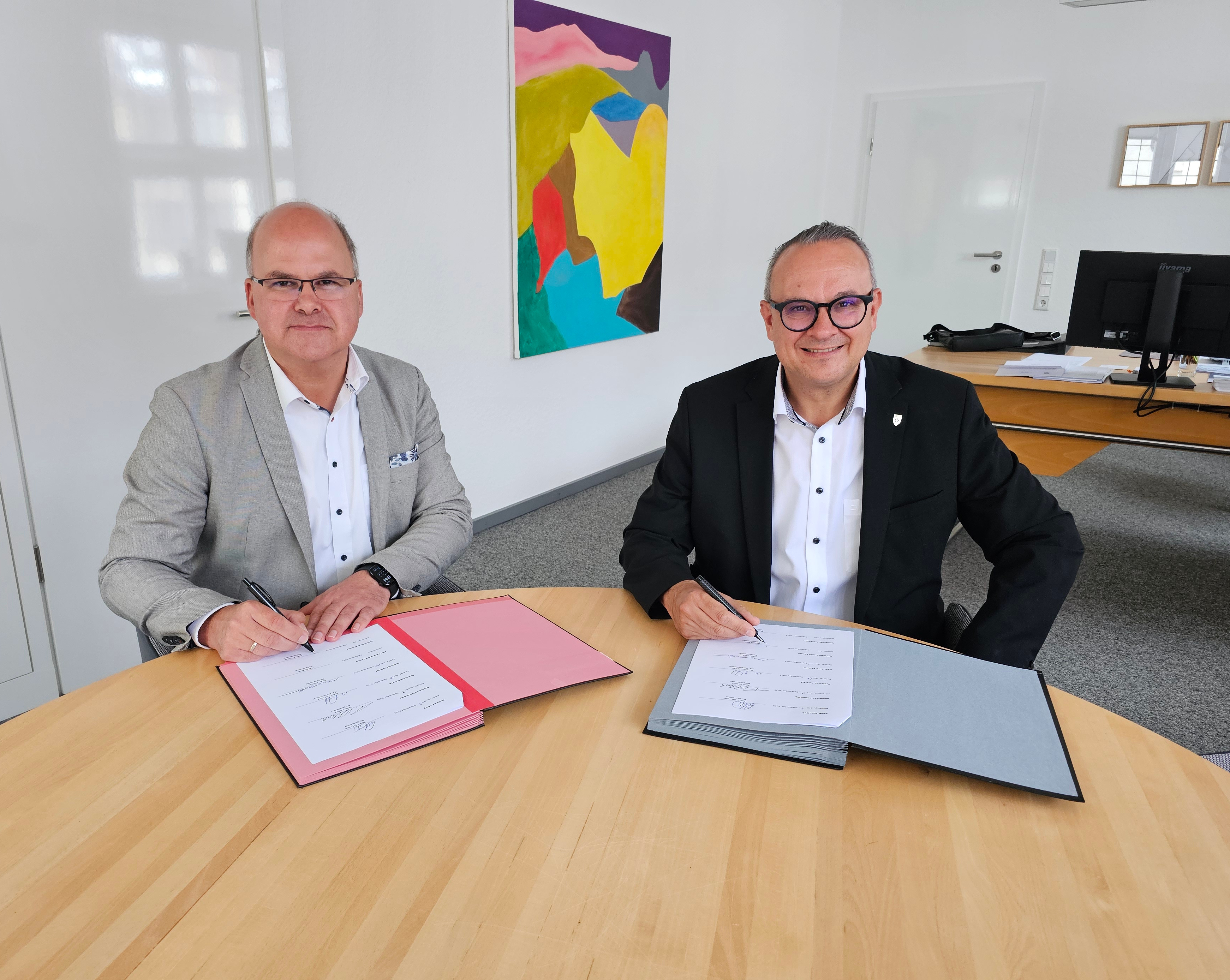 Die beiden Bürgermeister sitzen an einem Tisch. Vor ihnen liegen die zu unterzeichnenden Dokumente.